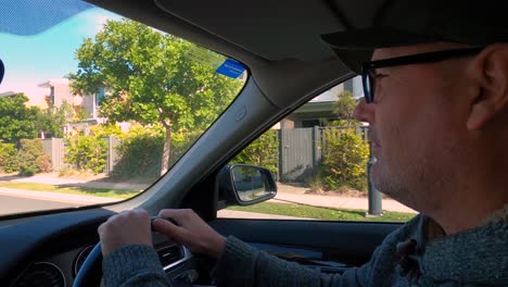 a mature man in glasses, casual clothes, and a cap, driving a car and talking