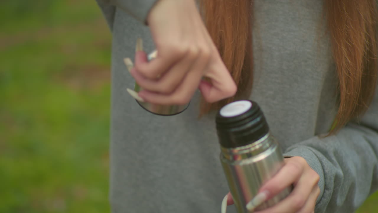 primer plano de una mujer joven con suéter gris desenroscando cuidadosamente la tapa del termo, presionando la cubierta interna para verter agua en la tapa en medio de un bosque verde sereno, con uñas delicadas y bien cuidadosas