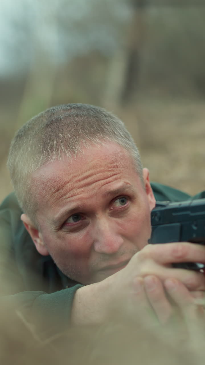 una vista de cerca de un hombre dentro de un agujero en el suelo en un área seca y cubierta de hierba, sosteniendo una pistola con un determinado y enfocado y mirando hacia atrás