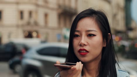 close up of the young pretty girl talking and recording a voice audio message on the smartphone in the city center