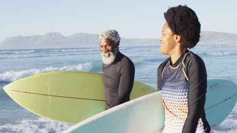 happy african american couple walking with surfboards on sunny beach