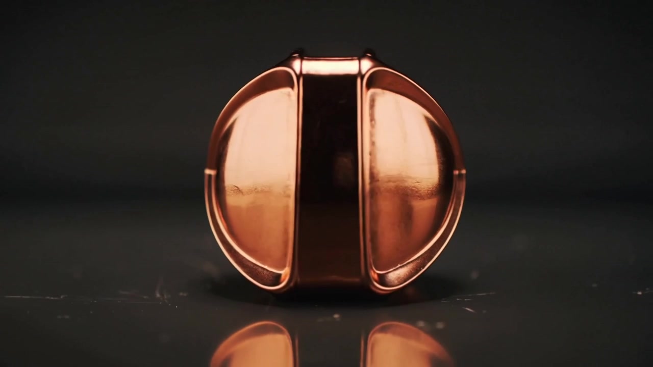Fidget geometrico in rame che si inclina e rotola, posandosi su un tavolo nero lucido mentre le luci dello studio lo illuminano