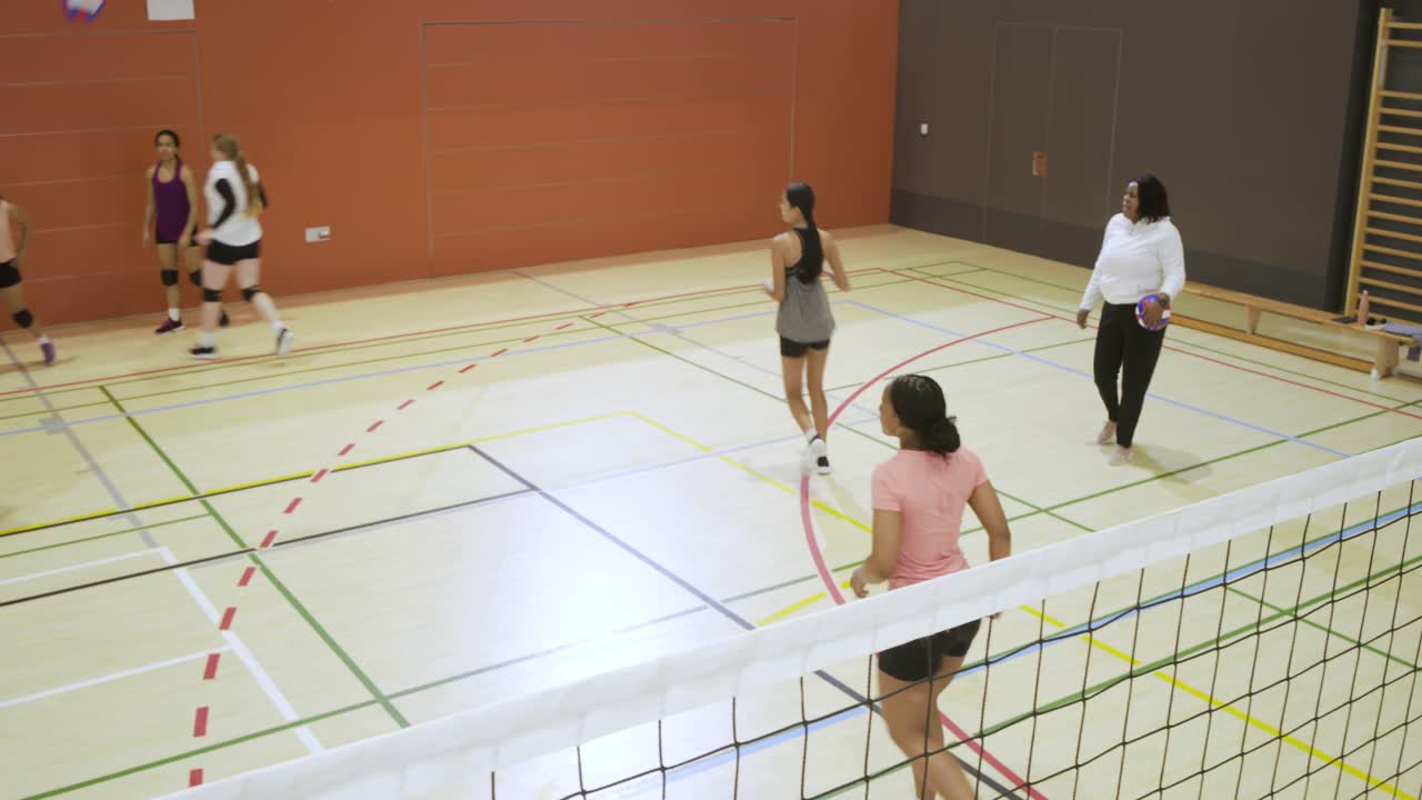 体育館でトレーニング中の多様な女性チーム。白いユニフォームの選手が紫色のボールをトスしてサーブを打ち、コーチが複数のボールを抱えている。