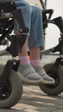 legs of little girl with multiple sclerosis sitting in wheelchair on walk with mother on street. woman pushes medical transport with daughter closeup