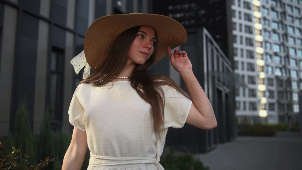 Una giovane donna con un abito bianco svolazzante e un cappello a tesa larga cammina a piedi nudi accanto a siepi curate vicino a un edificio moderno, con la calda luce del giorno che crea ombre morbide.