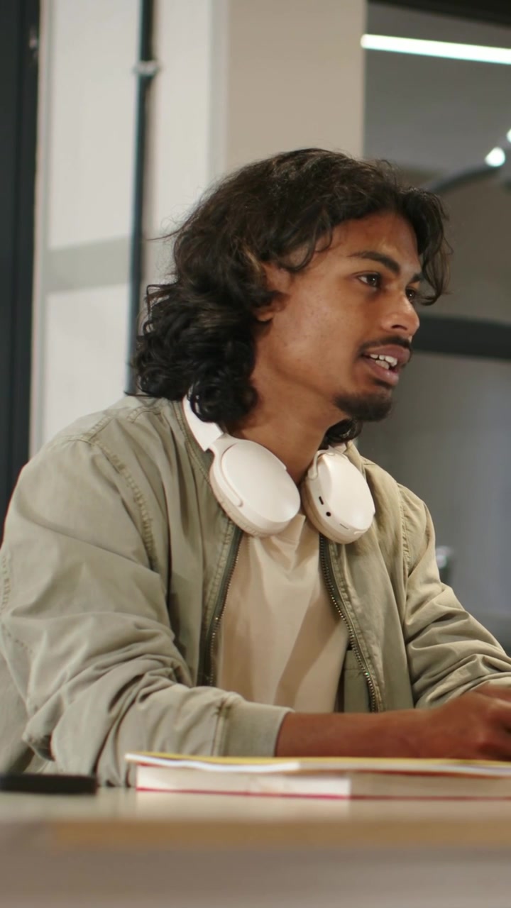 Vertical video: Turning man in green jacket and white headphones answering off-camera voice at work