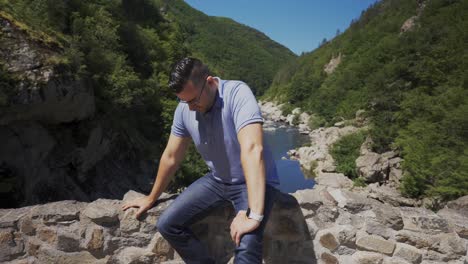 tourist sitting on the side railings of the devil's bridge, located right above the beautiful arda river, located next to the rhodope mountains in ardino, bulgaria