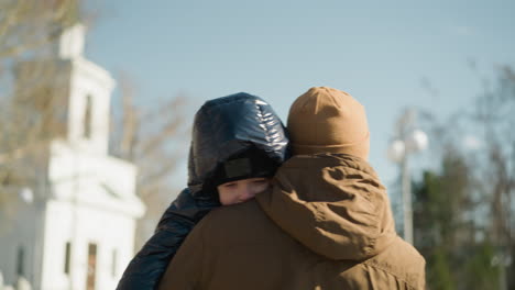 a close-up of father carrying his son and walking outdoors ,the father is dressed in a brown jacket and beanie, and the child in a black puffy jacket, with blurred view of a building