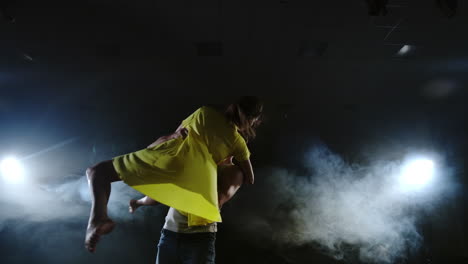 La-Cámara-Hace-Zoom-Y-Dispara-Desde-El-Plano-Completo-Al-Plano-Medio-En-Cámara-Lenta,-Una-Joven-Con-Un-Vestido-Amarillo-Y-Un-Hombre-Con-Una-Camisa-Blanca-Bailando-En-El-Escenario-Con-Focos-Y-Humo