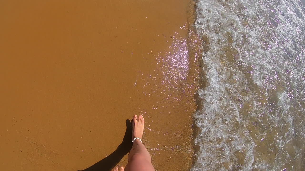 Woman filming her legs and feet as she is walking on golden, orange sand and through the ocean at the beach in malta.
