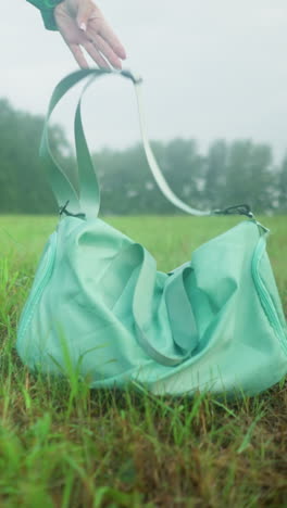 vista inferior de una persona con botas de lluvia amarillas y leggings dejando caer una bolsa de color menta en un campo cubierto de hierba, el fondo presenta un amplio campo de hierba y árboles bajo un cielo nublado