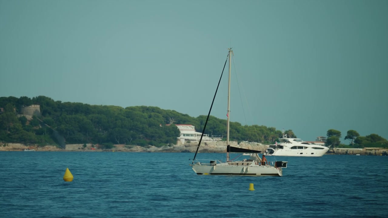 Barca a vela sull'acqua durante l'estate, con uno yacht di lusso e case costiere sullo sfondo sotto un cielo azzurro, Antibes, Francia