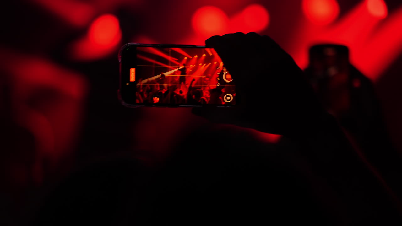 Enthusiastic crowd at a concert captures electrifying moments on smartphones, showcasing vibrant red and white stage lighting and live music atmosphere