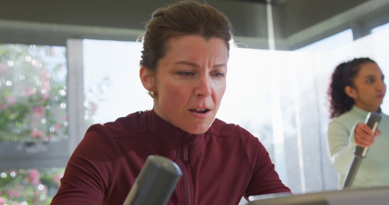 Diverse female friends, woman in burgundy top checking console for stats, pedaling at gym machine
