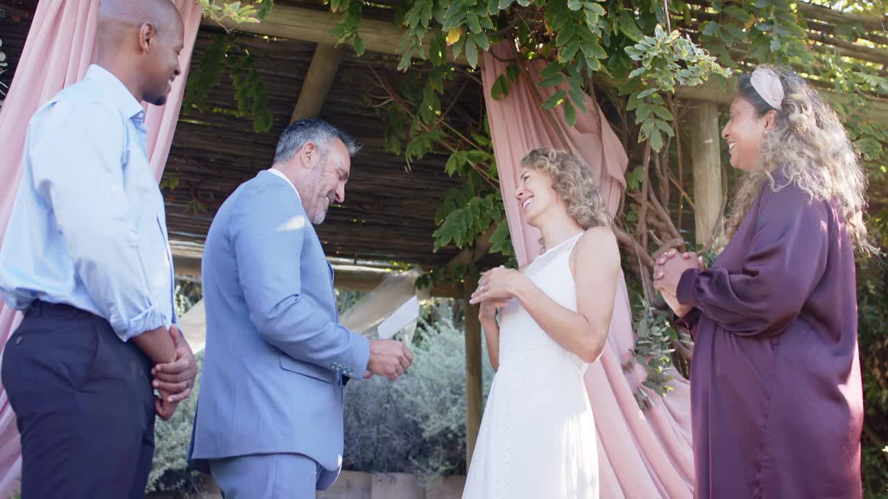Diverse wedding party watching suited groom reading vows and placing ring on bride beneath pergola