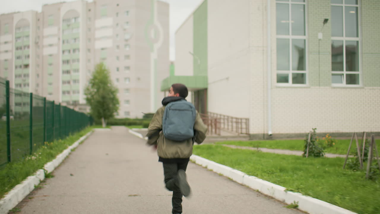 Vista trasera de un estudiante con mochila corriendo hacia el aula por un camino pavimentado bordeado de vegetación, vallas de hierro y altos edificios residenciales al fondo bajo un cielo nublado