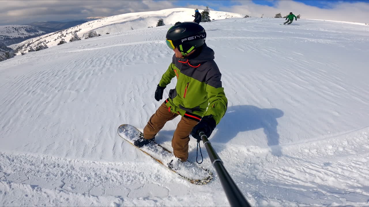Male snowboarder going down the hill takes selfie. Amazing scenery of the picturesque snowy mountains at backdrop.