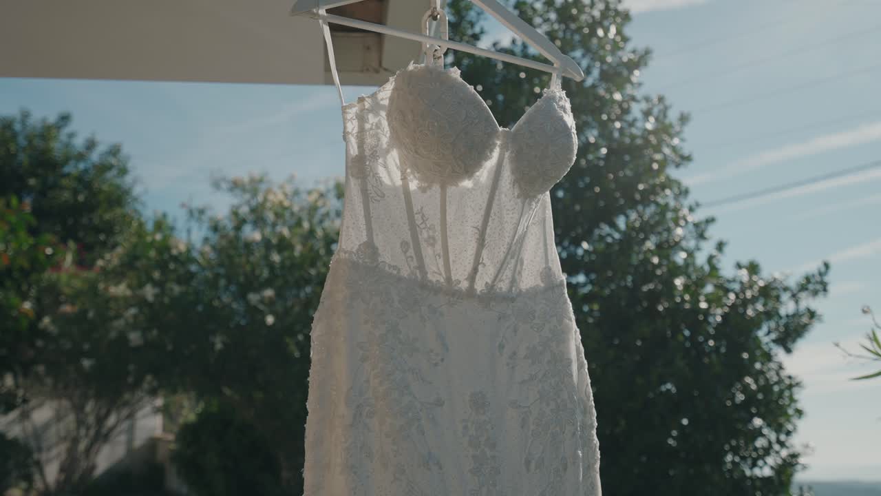 아침 햇살에 비치는 흰색 레이스 웨딩드레스가 옷걸이에 걸려 있고, 배경에는 나무와 하늘이 있어요.