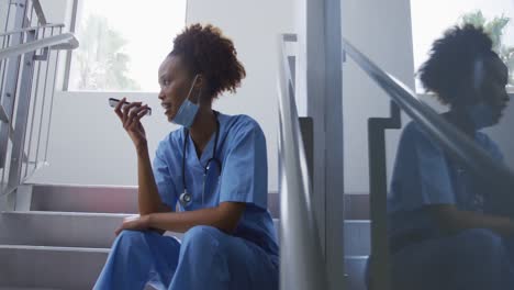 mixed race female doctor wearing face mask sitting on stairs in hospital talking on smartphone