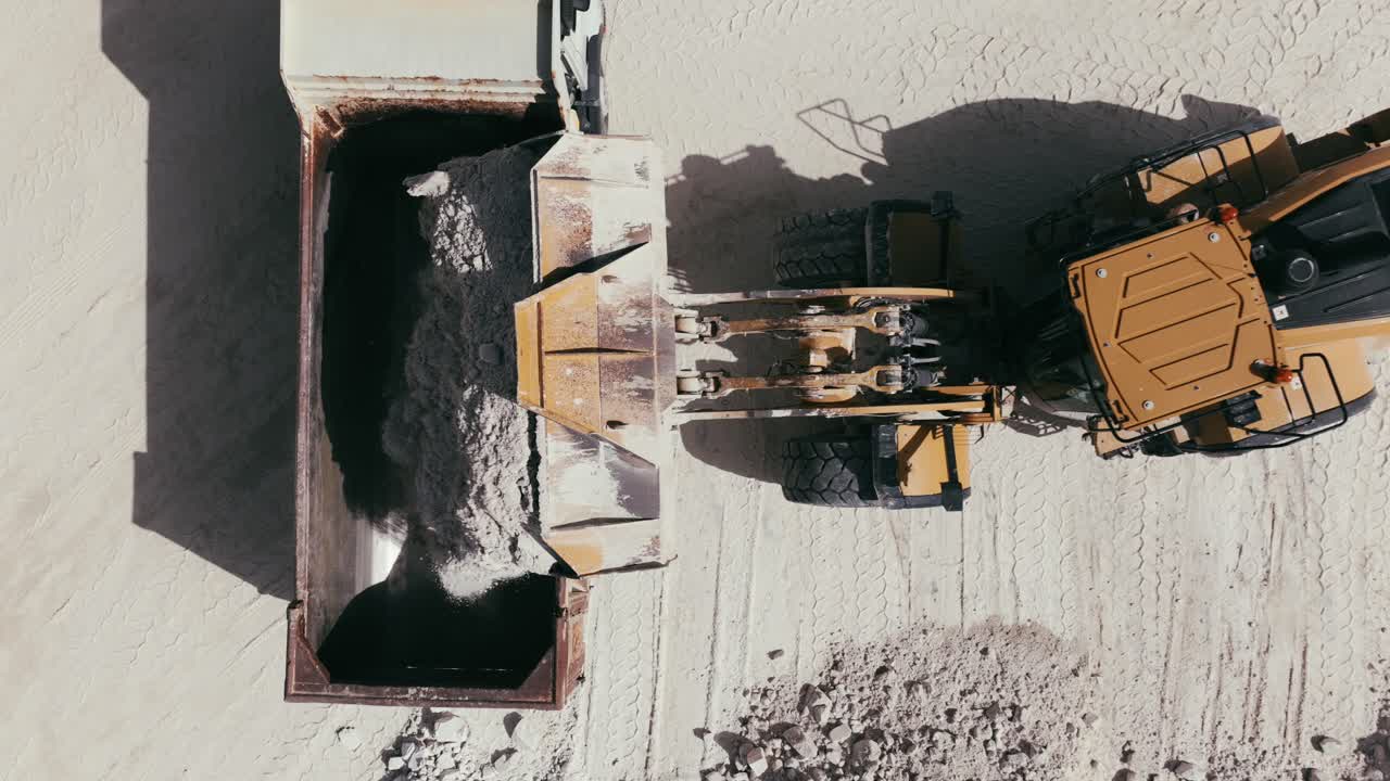 A top-down view of a heavy-duty loader in action, moving materials across an industrial site. The machine's large bucket is filled with earth or minerals. Aerial view.