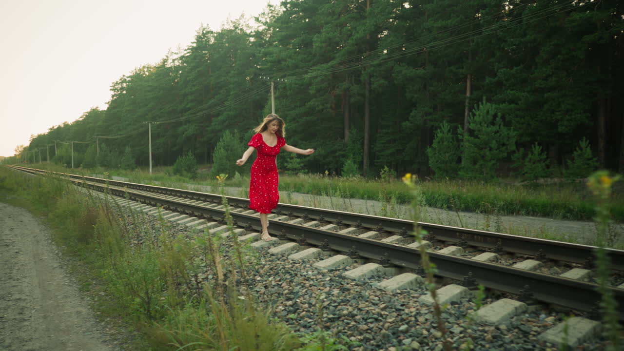 Side view of young woman in red dress running cautiously on rail sleeper through countryside area, surrounded by gravel, green vegetation, tall trees, and utility pole under soft natural daylight