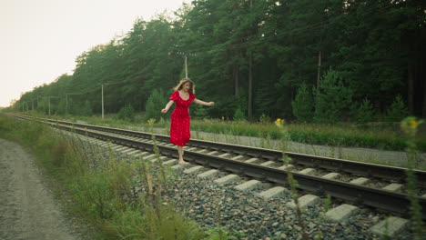 side view of young woman in red dress running cautiously on rail sleeper through countryside area, surrounded by gravel, green vegetation, tall trees, and utility pole under soft natural daylight