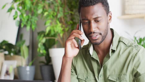 happy african american man sitting at table in kitchen, talking on smartphone