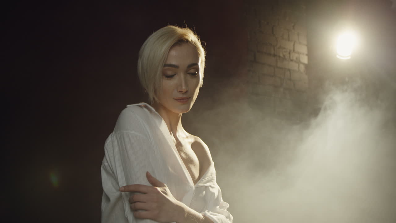 사진사가 그녀의 사진을 찍는 동안 연기와 함께 어두운 스튜디오에서 포즈를 취하는 백인 젊은 여성 모델의 클로즈업 보기