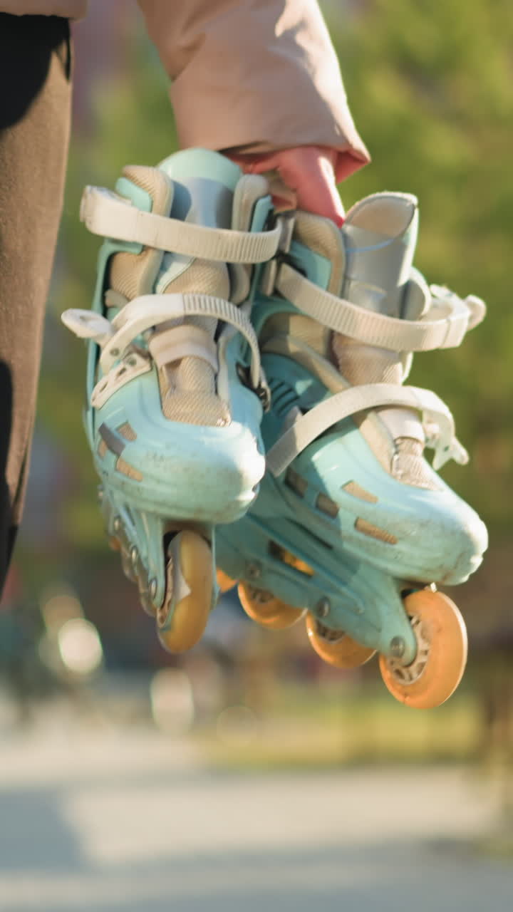 Front shot of a person holding a pair of rollerblades, wearing black trousers and a peach jacket, walking through a sunlit park with a blurred background of trees. The person's face is not visible