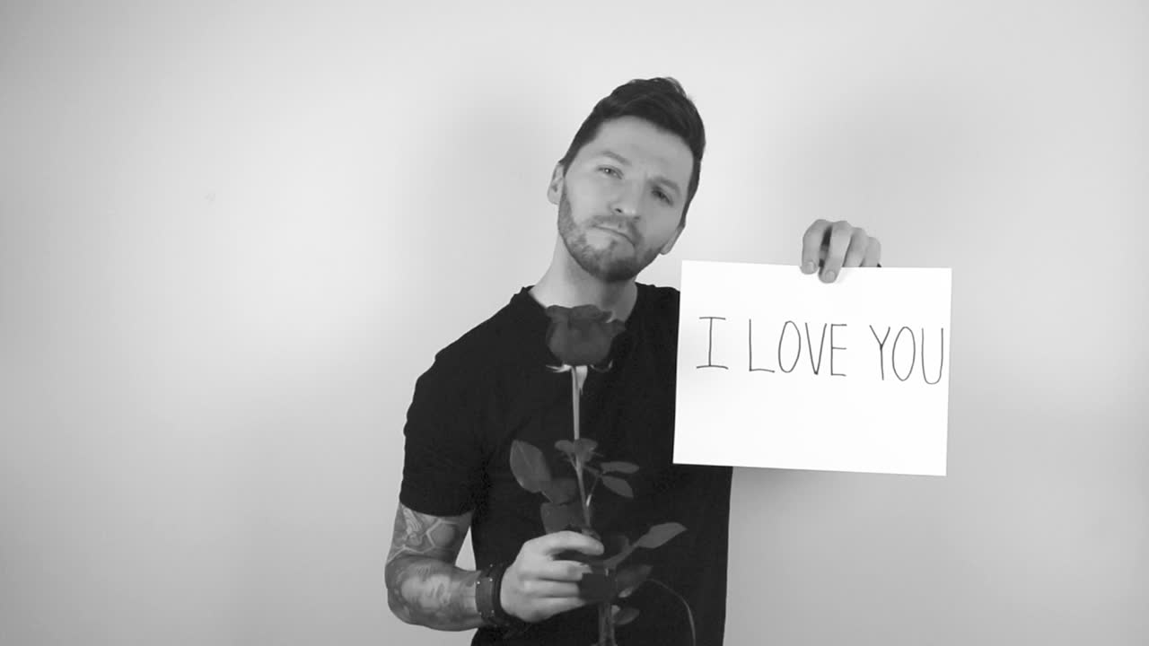 Grayscale footage of a tattooed man lifting up a blossoming rose flower and a sign that says I love you on a white paper while smiling and being in a good mood. Man on a white studio background.