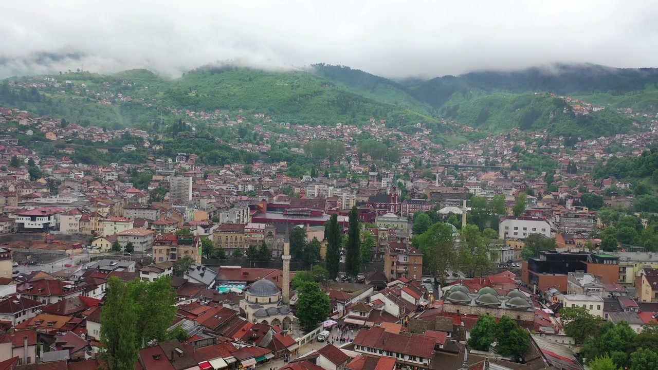 Cảnh quay drone trên cao góc rộng cho thấy sự mở rộng đô thị của Sarajevo, nổi bật với những mái nhà đỏ, các tòa nhà cao tầng hiện đại và những đám mây thấp bao phủ các ngọn núi xa ở Bosnia và Herzegovina