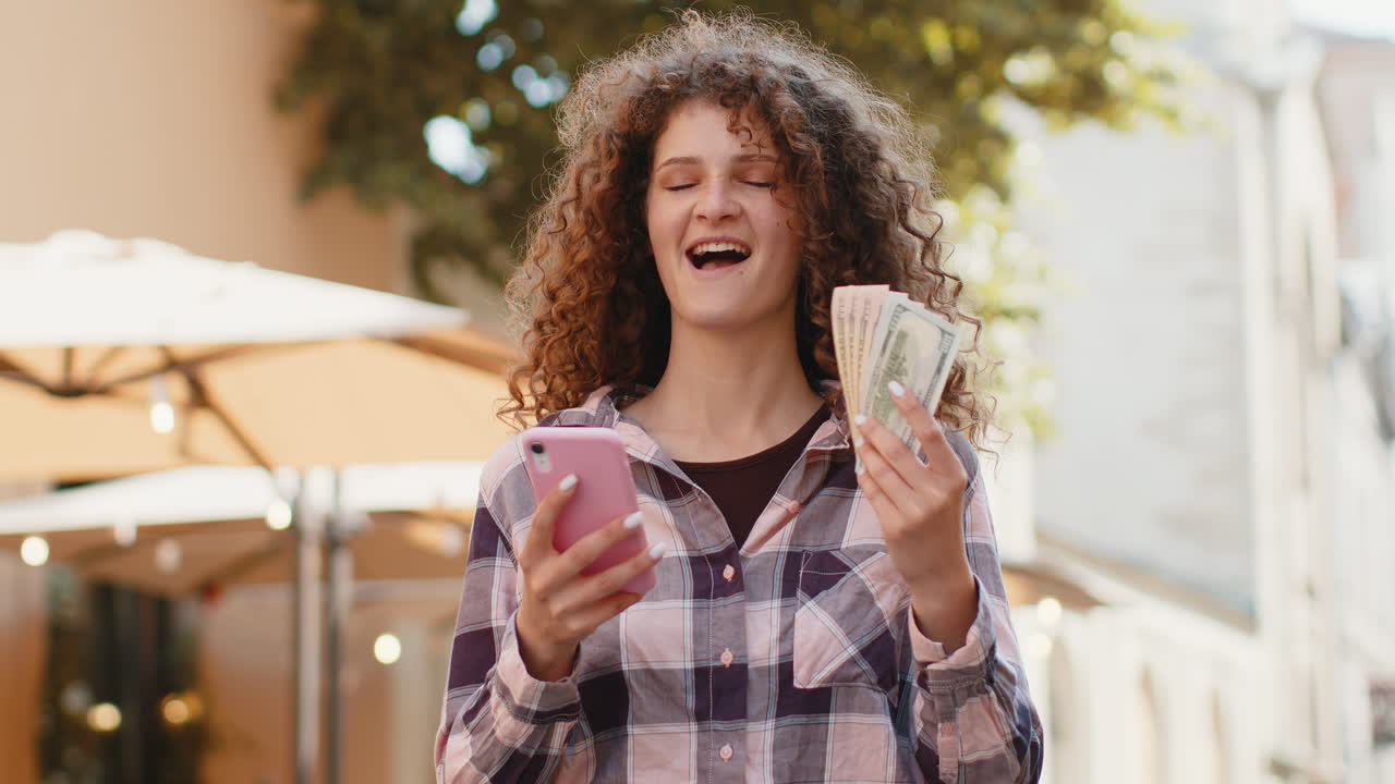 Happy young woman counting money dollar cash use smartphone calculator app in urban city street