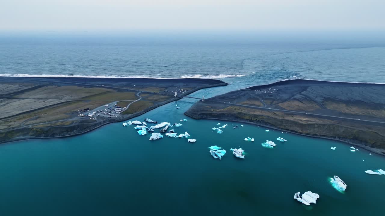Panoramica aerea verso lo sbocco di una laguna glaciale dove l'acqua di fusione turchese incontra l'Atlantico, incorniciata da lingue di sabbia nera e iceberg sparsi sotto un orizzonte pallido