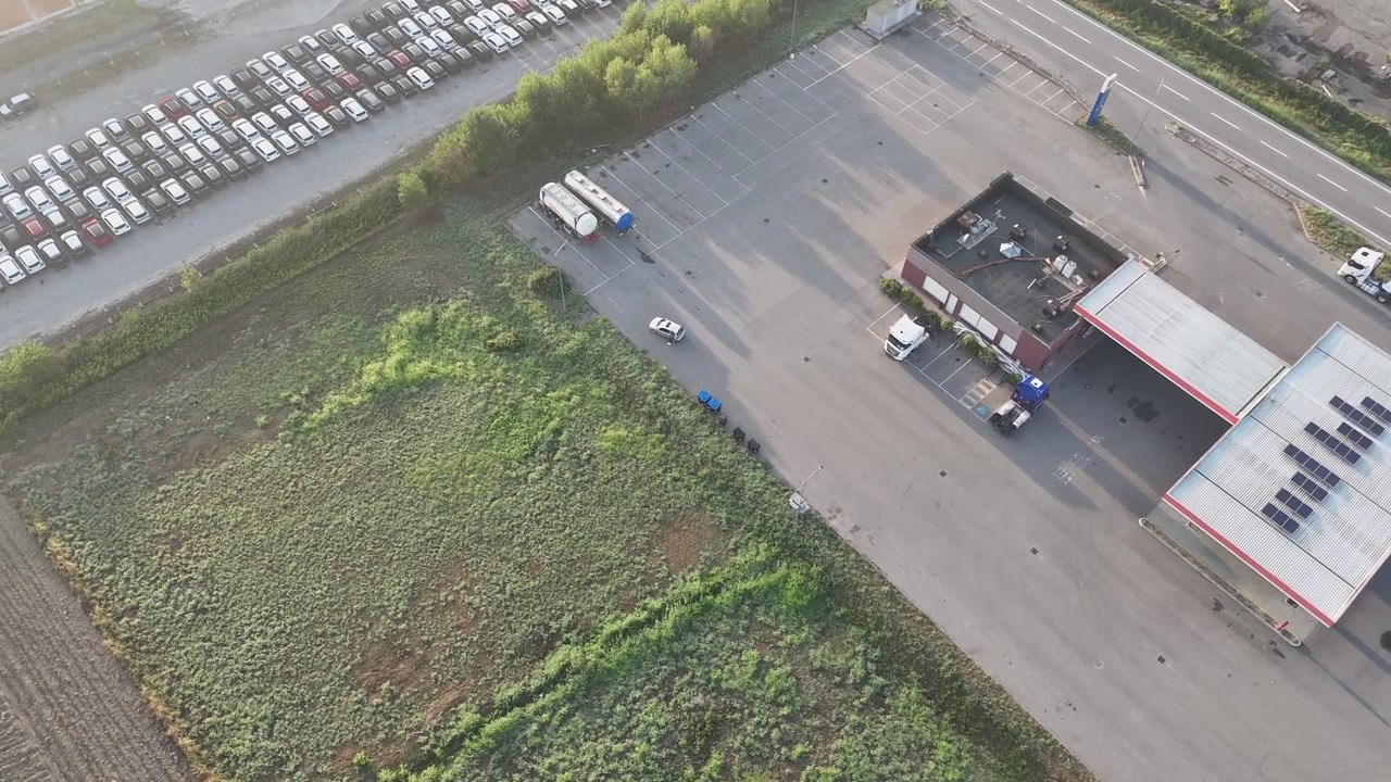 Fuel station with canopy, forecourt and roadside access stands beside industrial buildings, vehicle storage and cultivated fields during a drone descent.