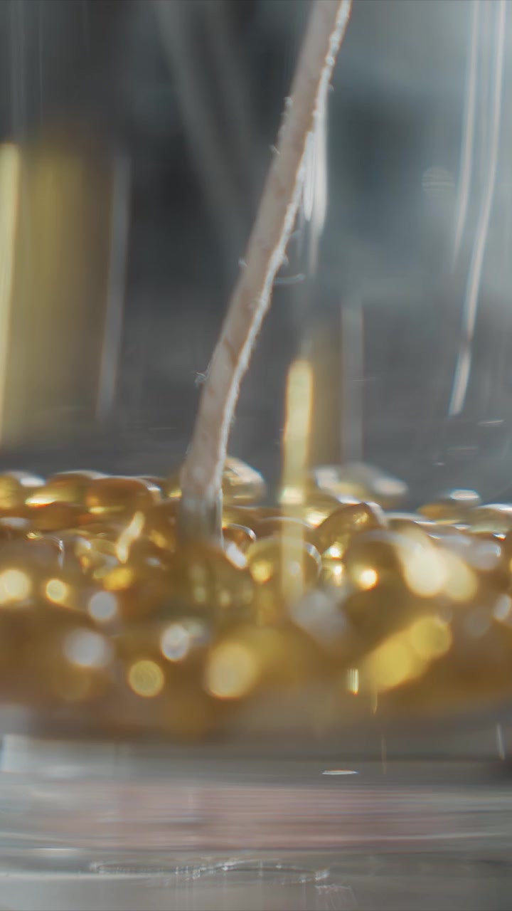 Primo piano di liquido dorato che scorre attraverso un tubicino sottile in un contenitore trasparente, creando bolle e particelle scintillanti. Il liquido appare viscoso con riflessi di luce calda su uno sfondo scuro.