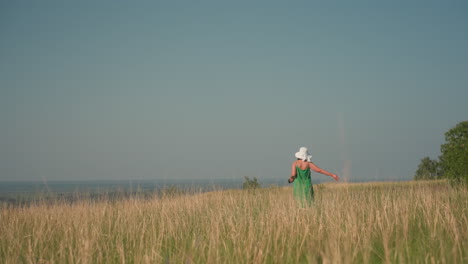 elegant woman in white floppy hat and green sundress walking slowly through golden tall grass field on sunlit hillside with camera hanging from hand, summer breeze rustling stalks