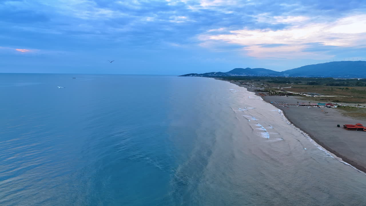 Seagulls fly over the vast blue tranquil waterscape of the Adriatic Sea at sunset. View on the beach with tourist infrastructure in Montenegro from drone