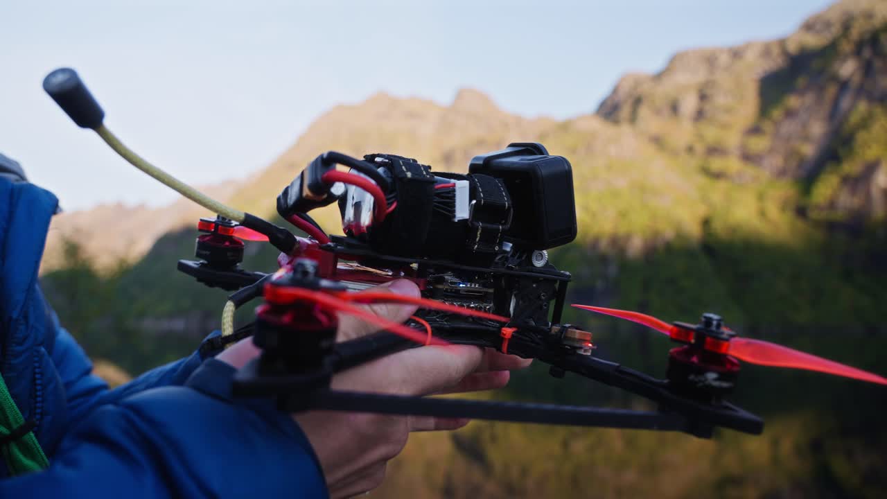 A person adjusts a red racing drone outdoors near water in Vestarelen, Norway, during sunrise.