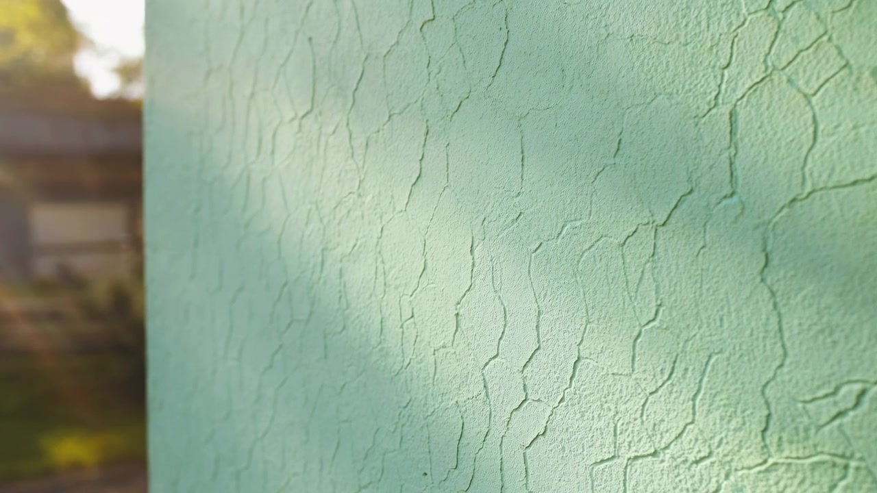 Oblique close view of mint green exterior wall with peeling paint, rough stucco texture, irregular cracks, and weathered surface lit by warm sunlight, emphasizing aging, repair needs, and building