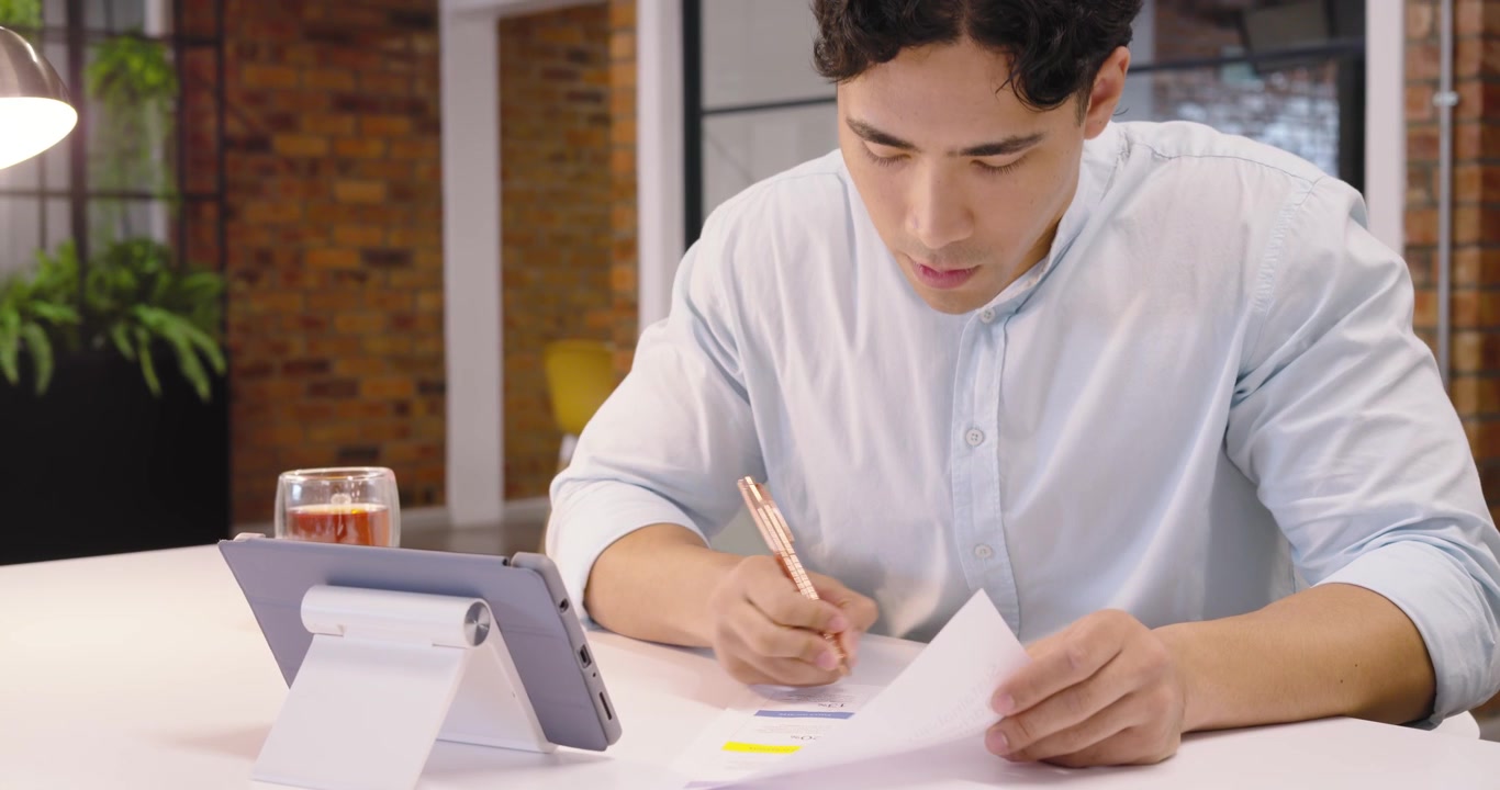 Pria Asia dewasa melirik antara tablet dan grafik cetak, menganotasi laporan dengan pena di meja