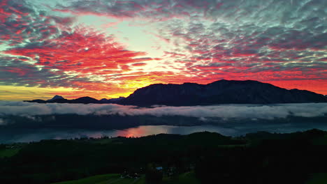 Montañas-épicas-De-Puesta-De-Sol-De-Fuego-Y-Capa-De-Niebla-Sobre-El-Lago,-Fondo-Aéreo-Panorámico-Que-Establece