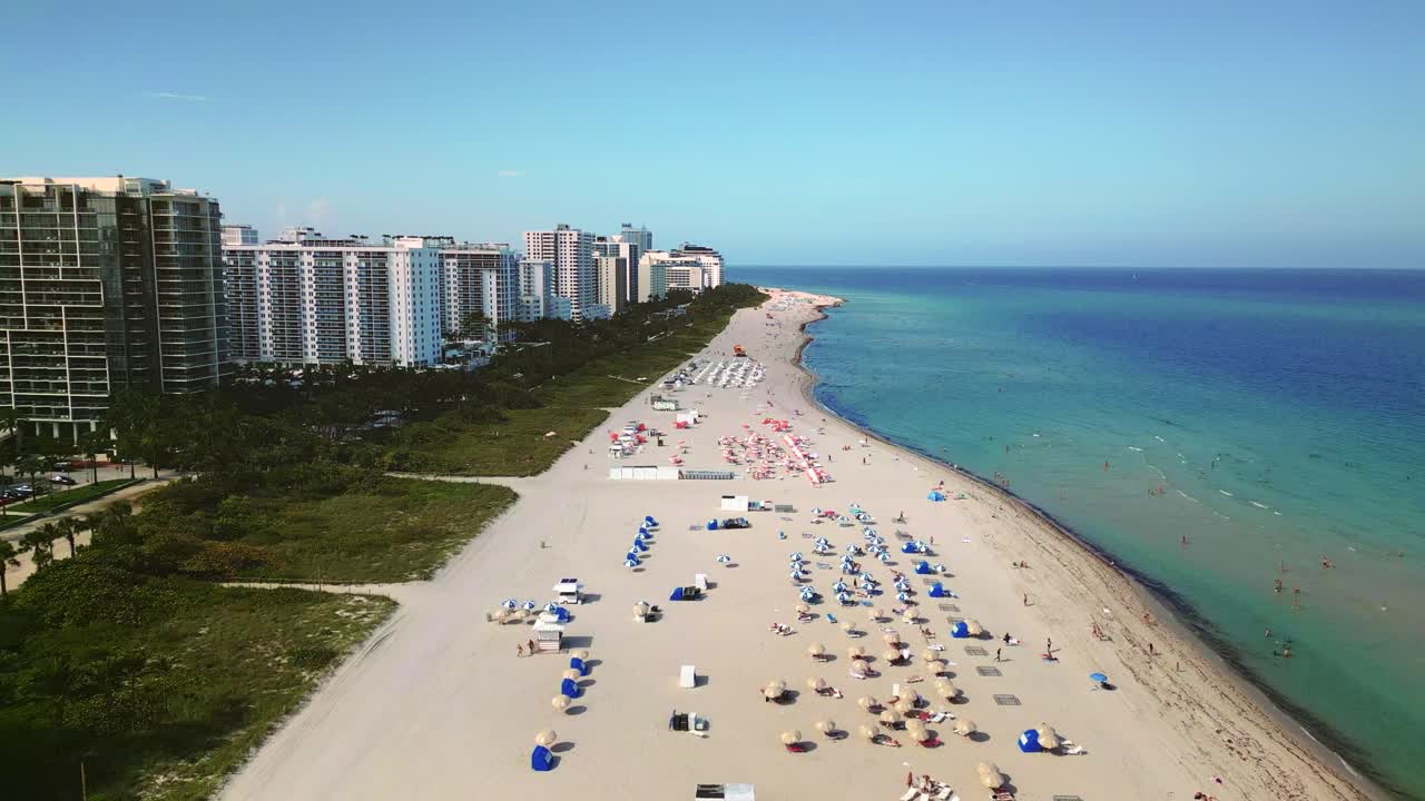 Aerial 4K view of Miami's Iconic South Beach, soaring over sunbeds and umbrellas as beachgoers relax along the pristine white sands, framed by turquoise waters and luxury resorts.