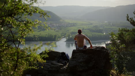 Hiker-taking-break-above-Danube-river,-wide-cinematic-shot,-austria,-durnstein,-europe,-hazy-mountain-valley-landscape,-independent-male-traveller,-sitting-down,-bare-chest,-hike-trail-wandering