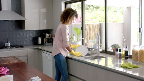 happy biracial woman washing up dishes at sink in sunny kitchen, copy space, slow motion