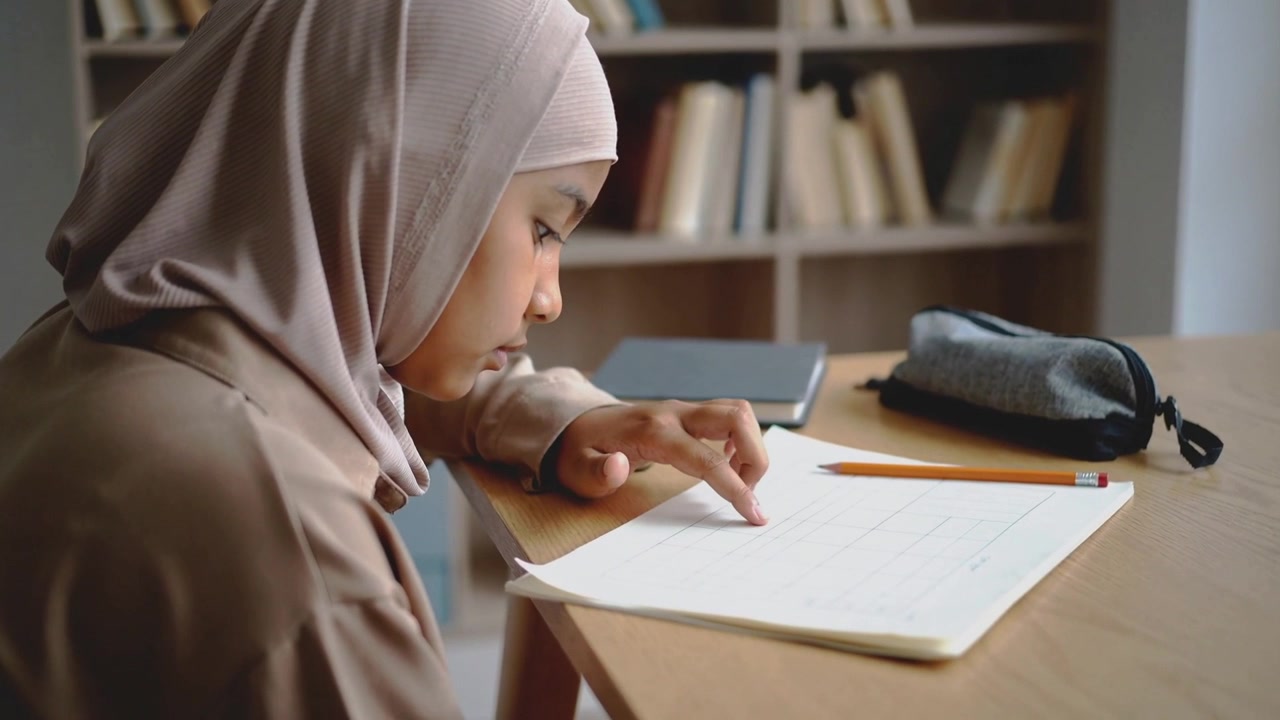 Seorang gadis membungkuk, mengenakan syal dan lengan panjang, mengikuti baris-baris buku kerja untuk mengerjakan pekerjaan rumah di rumah, dengan pensil.