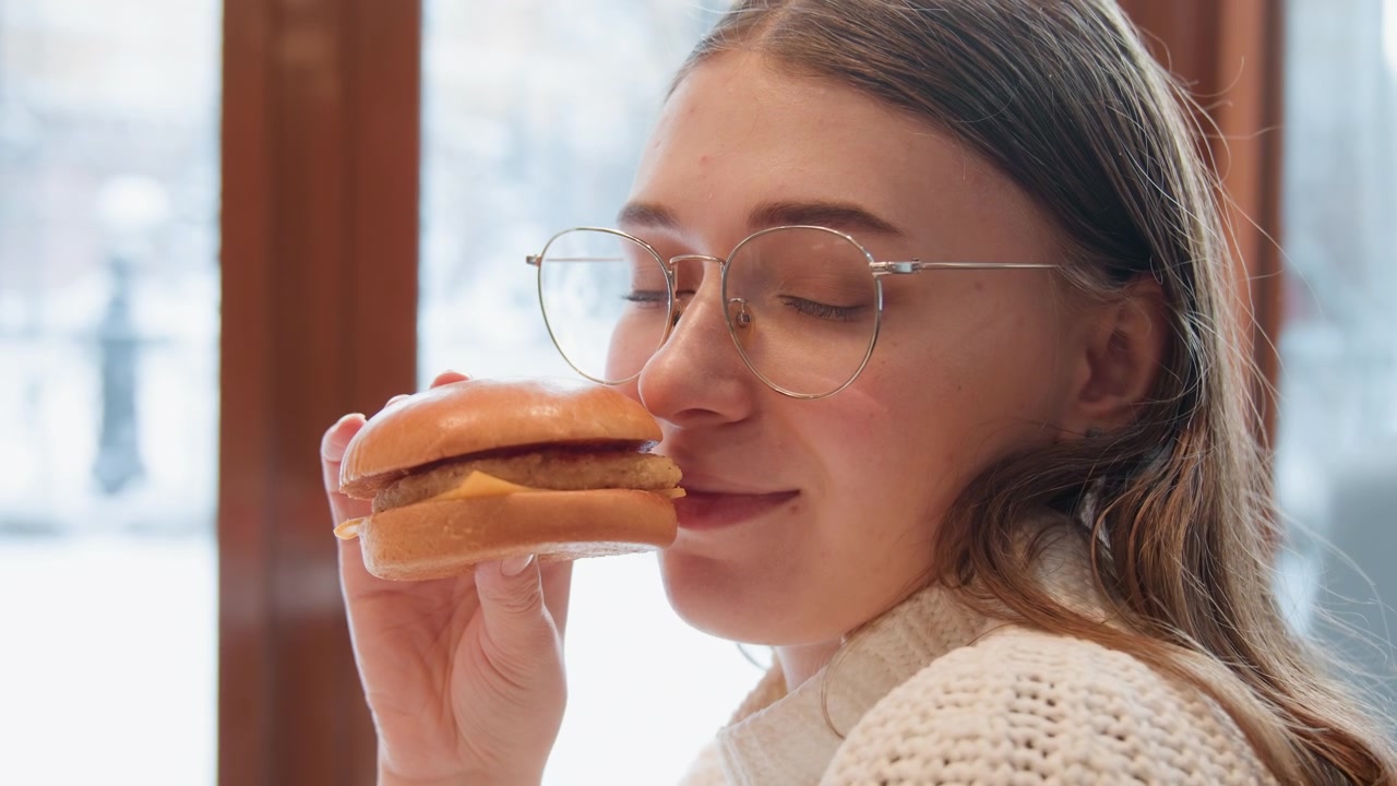 Young woman indoors near snowy window lifts sandwich toward mouth, smiling at camera with subtle head turn, gentle hand motion, soft winter light, and cozy room atmosphere during relaxed food moment
