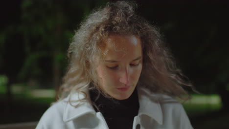 young girl seated outdoors at night looking down in deep reflection with curly hair gently swaying in breeze under soft ambient light, surrounded by dark background of trees and nature