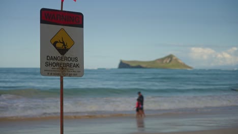 Ein-Langsamer-Schwenk-über-Das-Gefährliche-Shorebreak-Warnschild-Mit-Einem-Elternteil,-Der-Mit-Seinen-Kindern-Spielt