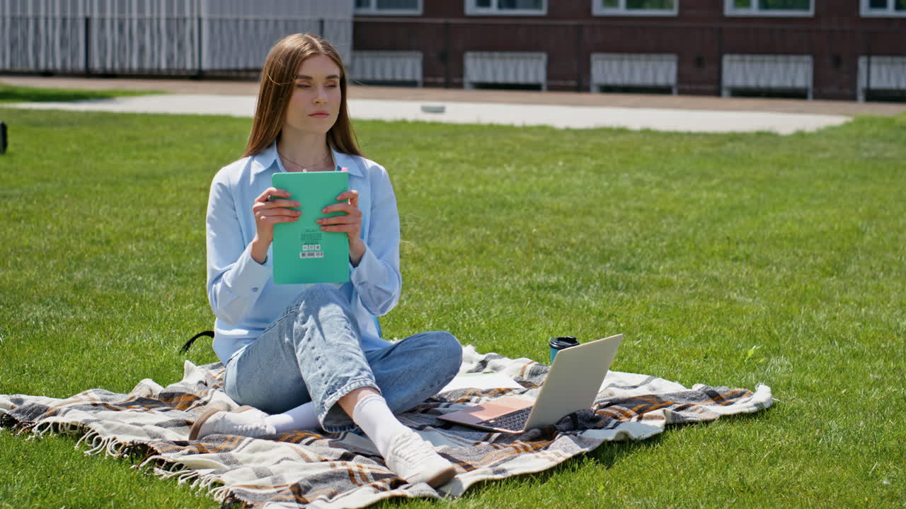 Thoughtful student writing notes on grassy campus sunny day. Young girl learning