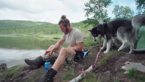 man and dog resting by the lake preparing a single burner portable camping stove while trekking in ånderdalen national park in the island of senja, norway